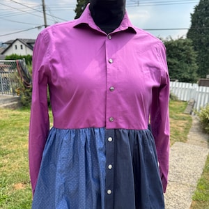 May include: A two-tone shirt with a purple top and a blue skirt-like bottom. The top has long sleeves and a button-down front. The bottom is a combination of solid and polka-dot blue fabrics. The shirt is displayed on a mannequin.