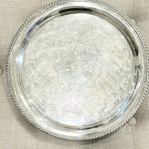 Może przedstawiać: Okrągła, srebrna taca do serwowania z ozdobnym, wytłoczonym wzorem kwiatowym. Taca ma podniesioną krawędź z wzorem w kształcie liny. Taca jest prawdopodobnie wykonana z metalu i nadaje się do serwowania potraw lub eksponowania przedmiotów.