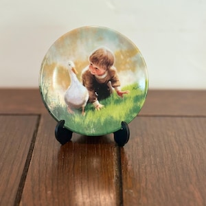 Puede incluir: Un plato decorativo con un niño y un pato en un campo de hierba. El plato tiene un diseño pintado con un fondo verde claro y amarillo. El plato se exhibe en un soporte negro.