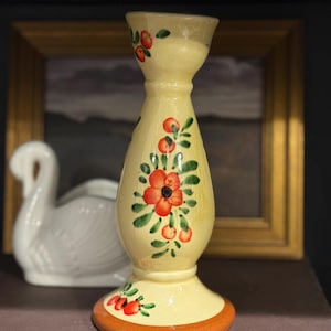 Puede incluir: Un candelero de cerámica amarilla con flores rojas y hojas verdes pintadas a mano. La base tiene un borde naranja. El candelero es un objeto decorativo.