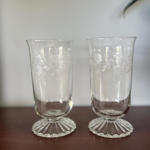 May include: A pair of clear glass footed tumblers featuring a delicate etched floral vine pattern around the upper body. Each glass has a smooth cylindrical bowl that transitions into a short stem and a wide, circular base with radial ribbing.