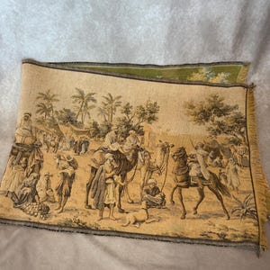 Puede incluir: Un tapiz tejido rectangular que muestra una escena del desierto con figuras montadas en camellos y caballos, palmeras y tiendas de campaña en una paleta de colores beige y marrón. La tela tiene un borde con flecos en el lado derecho.