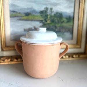 Puede incluir: Una vasija de cerámica color terracota con dos asas laterales y una tapa blanca lisa. El recipiente presenta un cuerpo redondeado y un acabado mate en el exterior, mientras que la tapa tiene un esmalte blanco brillante y un pomo central.