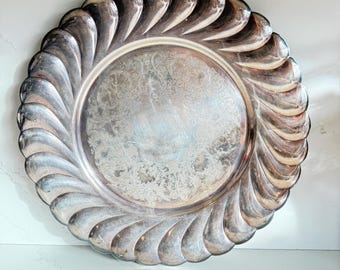 May include: Silver-toned serving tray with a decorative, swirling edge. The tray has a circular shape with an ornate, etched floral pattern in the center. The rim features a repeating, curved design, adding a vintage aesthetic.