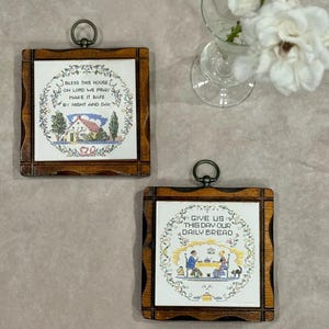 May include: Two framed ceramic tiles with religious text and imagery. The tiles have a dark wood frame and a metal ring for hanging. One tile reads "Bless this house" and depicts a house. The other tile reads "Give us this day our daily bread".