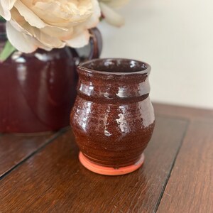 May include: A small, handmade ceramic vase with a rich, dark brown glaze. The vase has a rounded body and a slightly flared rim, sitting on a contrasting orange base. A larger burgundy vase and cream-colored flowers are in the background.