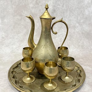 May include: A vintage brass tea set, including a tall, ornate pitcher with a curved spout and handle, a tray, and several small goblets. The set features etched floral patterns and a warm, golden hue.