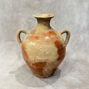 Puede incluir: Una ánfora antigua y desgastada con dos asas. El recipiente de cerámica presenta un color base beige con acentos marrón rojizo y un borde ligeramente ensanchado. Su aspecto envejecido sugiere importancia histórica, ideal para decoración.