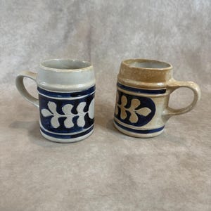 May include: Two ceramic mugs with handles, each featuring a cylindrical body and a raised horizontal band near the rim. One mug is off-white and the other is tan, both decorated with a dark blue oval containing a white leaf-like botanical pattern.