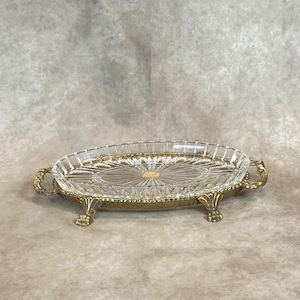 Puede incluir: Una bandeja ovalada de vidrio transparente con base y asas de metal dorado. El vidrio presenta un detallado patrón tallado. La bandeja se apoya en cuatro patas ornamentadas, realzando su estética vintage. Mide unos 25 cm de largo.