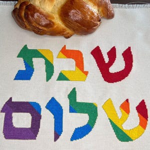 Peut inclure: Un tissu blanc à bord frangé orné d'une inscription hébraïque en point de croix aux couleurs arc-en-ciel vives. Les lettres présentent des dégradés de rouge, orange, jaune, vert, bleu et violet. Un pain tressé est posé sur le haut du tissu.