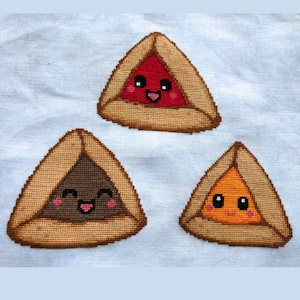 May include: Three triangular cross-stitch patches featuring cute, smiling faces. One patch has a red center, one has a dark brown center, and one has an orange center, all framed by a tan, textured border on a light blue fabric background.