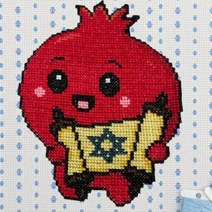 May include: A needlepoint canvas featuring a red pomegranate character holding a yellow scroll with a blue Star of David. The background is white with a repeating blue hamsa pattern. Two spools of light blue embroidery thread sit at the bottom.