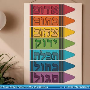 May include: A cross-stitch pattern featuring seven crayons in various colors: red, orange, yellow, green, teal, blue, and purple. Each crayon displays a word in Hebrew. The pattern is on a cream-colored background.