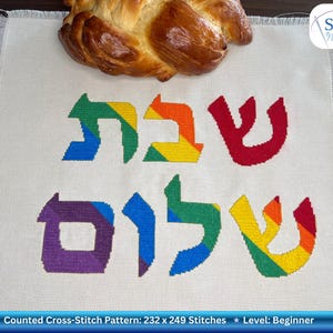 May include: A cross-stitch pattern featuring rainbow Hebrew lettering and a challah loaf. The text says "Shabbat Shalom." The pattern is for beginners and measures 59 x 63 cm. A Shalom Needlecraft logo is in the upper right corner.