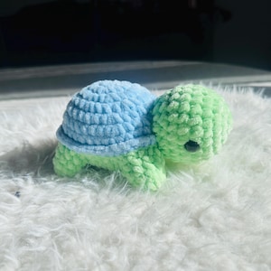 May include: A handmade crochet turtle in shades of blue and green. The turtle has a light blue shell and a green head, legs, and body. The turtle has a black eye and is sitting on a white, fluffy surface.