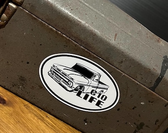 May include: An oval sticker featuring a black and white illustration of a classic pickup truck. The text "C10 LIFE" is printed below the truck. The sticker is affixed to a weathered, dark metal surface.