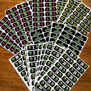 May include: A collection of black rectangular stickers with various designs. Some feature a green frog mascot with the word "Eckers", others have pink ribbons, and some display numbers like 2, 23, 24, and 28. The stickers are arranged on a wooden surface.