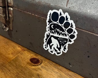 May include: A black and white sticker in the shape of a Bigfoot footprint. The footprint design includes a silhouette of Bigfoot, trees, mountains, and a crescent moon. The sticker is adhered to a gray metal toolbox.