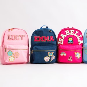 May include: Four personalised backpacks in pink, blue, and denim blue. Each backpack has a name in a decorative font, with patches of flowers, hearts, and other designs. The backpacks have gold zips and are designed for school.