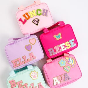 May include: Five colorful lunch bags with handles. Each bag is a different pastel color and has a name or word spelled out in glittery letters. The bags are decorated with patches of hearts, a paw print, a butterfly, a diamond, and a unicorn.
