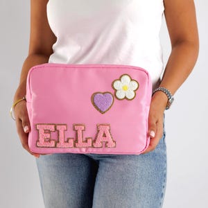 May include: A pink rectangular cosmetic bag with the name "ELLA" in pink varsity letters. The bag has a purple heart and a white flower with a yellow center. The bag is held by a person wearing a white t-shirt and blue jeans.