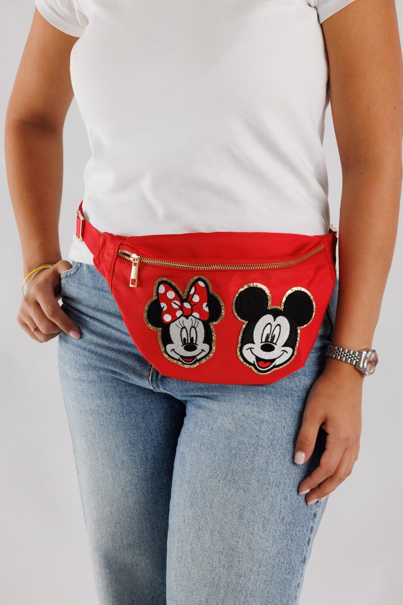 May include: A red fanny pack featuring embroidered images of Minnie Mouse and Mickey Mouse. The pack has a gold zipper and a red adjustable strap. The characters are outlined in gold and have black and white details.