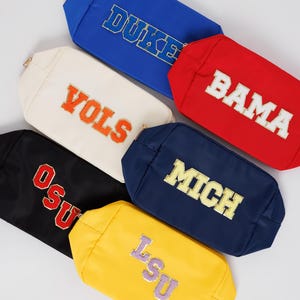 May include: A collection of rectangular cosmetic bags in various colors, each embroidered with a different college team name. Colors include blue, red, black, navy, yellow, and cream. The bags have gold zippers and are arranged on a white background.