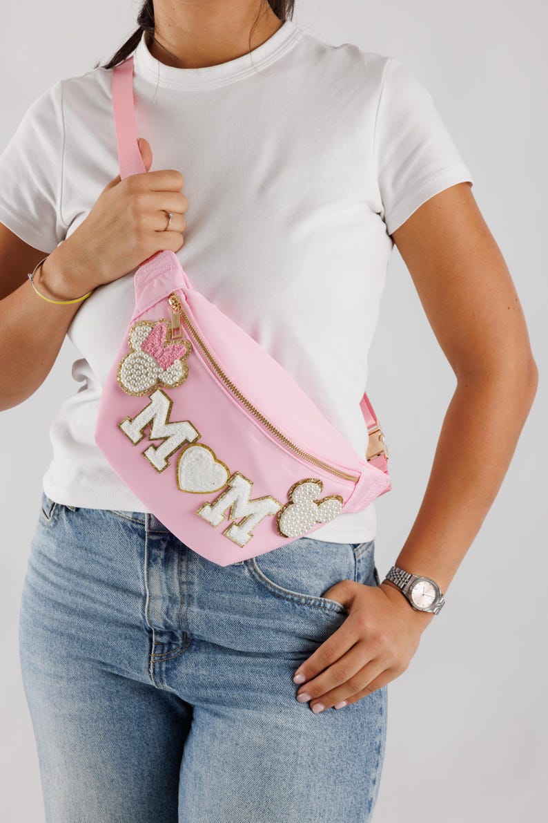 May include: Pink fanny pack with gold zipper and strap. The bag features the word "MOM" in white letters with a heart and pearl accents. A Minnie Mouse-inspired design is on the left side. The bag is worn across the body.