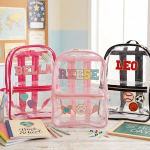 Puede incluir: Tres mochilas transparentes con los nombres "BELLA", "REESE" y "LEO". Las mochilas tienen detalles coloridos, incluyendo rayas rosas, negras y blancas, y parches de corazones, una huella de pata, una mariposa, una pelota de béisbol y un balón de fútbol.