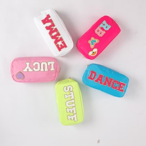 Può includere: Cinque astucci colorati con cerniera e testo ricamato. Gli astucci sono bianchi, rosa, rosa acceso, azzurro e verde neon. Il testo sugli astucci dice "EMMA", "RB", "LUCY", "DANCE" e "STUFF". Gli astucci hanno una forma rettangolare.