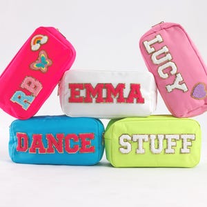 May include: A collection of colorful rectangular cosmetic bags. The bags are in pink, white, blue, and yellow-green. Each bag has a word or initials embroidered on it: "RB", "EMMA", "LUCY", "DANCE", and "STUFF". The "RB" bag has a rainbow and butterfly.