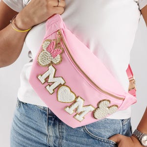 May include: Pink fanny pack with gold zipper and strap. The word "MOM" is spelled out in white letters with gold trim, and decorated with pearl and pink accents. A stylish accessory.