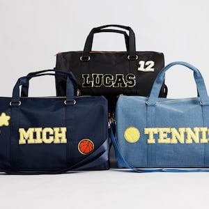Puede incluir: Tres bolsas de deporte personalizadas en azul marino, negro y azul vaquero. La bolsa azul marino tiene "MICH" con una pelota de baloncesto y una carita sonriente. La bolsa negra tiene "LUCAS" y el número 12. La bolsa vaquera tiene "TENNIS" con una pelota de tenis y un corazón.