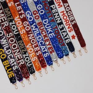 May include: A collection of sequined bag straps in various colors, each displaying a team name or slogan in white lettering. The straps have gold-colored clasps and include phrases like "GO BLUE" and "AGGIES".