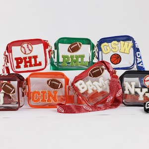 May include: A collection of clear, square crossbody bags with various sports-themed designs. Each bag features a different team's initials, a football or baseball, and a matching strap. The bags come in red, green, orange, blue, brown, and black.