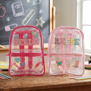 Può includere: Due zaini rosa trasparenti con i nomi "BELLA" e "REESE" in lettere glitterate. Ogni zaino ha un telaio rosa, spallacci e una tasca frontale con cerniera. Le toppe decorative includono cuori, farfalle e impronte di zampe. Su un tavolo di legno.