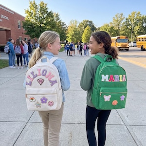 May include: Two girls walking on a sidewalk, each wearing a backpack. One backpack is cream-colored with the name "LILLONA" and decorative patches. The other is green with the name "MARIA" and patches. School buses are visible in the background.