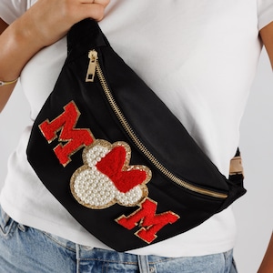 Könnte beinhalten: Schwarze Bauchtasche mit goldenem Reißverschluss und dem Wort "MOM" in Rot, mit Herz- und Perlenverzierungen. Die Tasche wird quer über dem Körper getragen. Die Tasche besteht aus schwarzem Material.