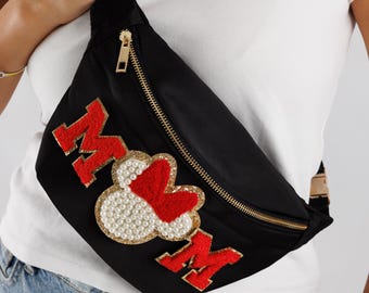 May include: Black fanny pack with gold zipper and the word "MOM" in red, with a heart and pearl embellishment. The bag is worn across the body. The bag is made of a black material.
