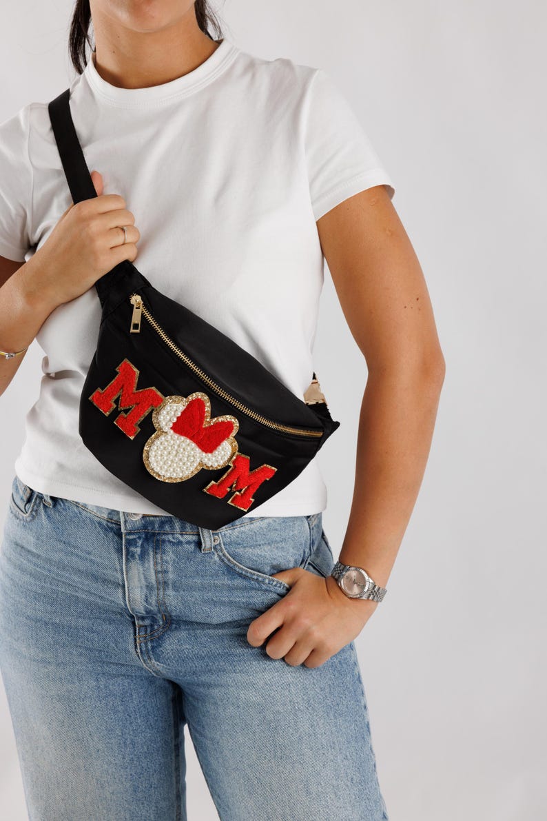May include: Black fanny pack with a gold zipper and a black strap. The pack features the word "MOM" in red letters with a pearl and red heart design in the center. A stylish accessory.