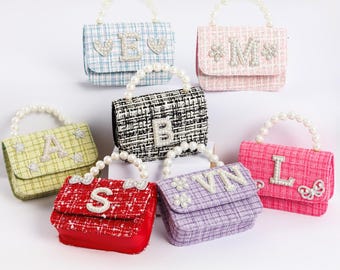 May include: A collection of small, colorful handbags with pearl handles. Each bag features a different initial or design in sparkling embellishments. The bags are in various colors, including blue, pink, red, and green, and have a textured fabric.