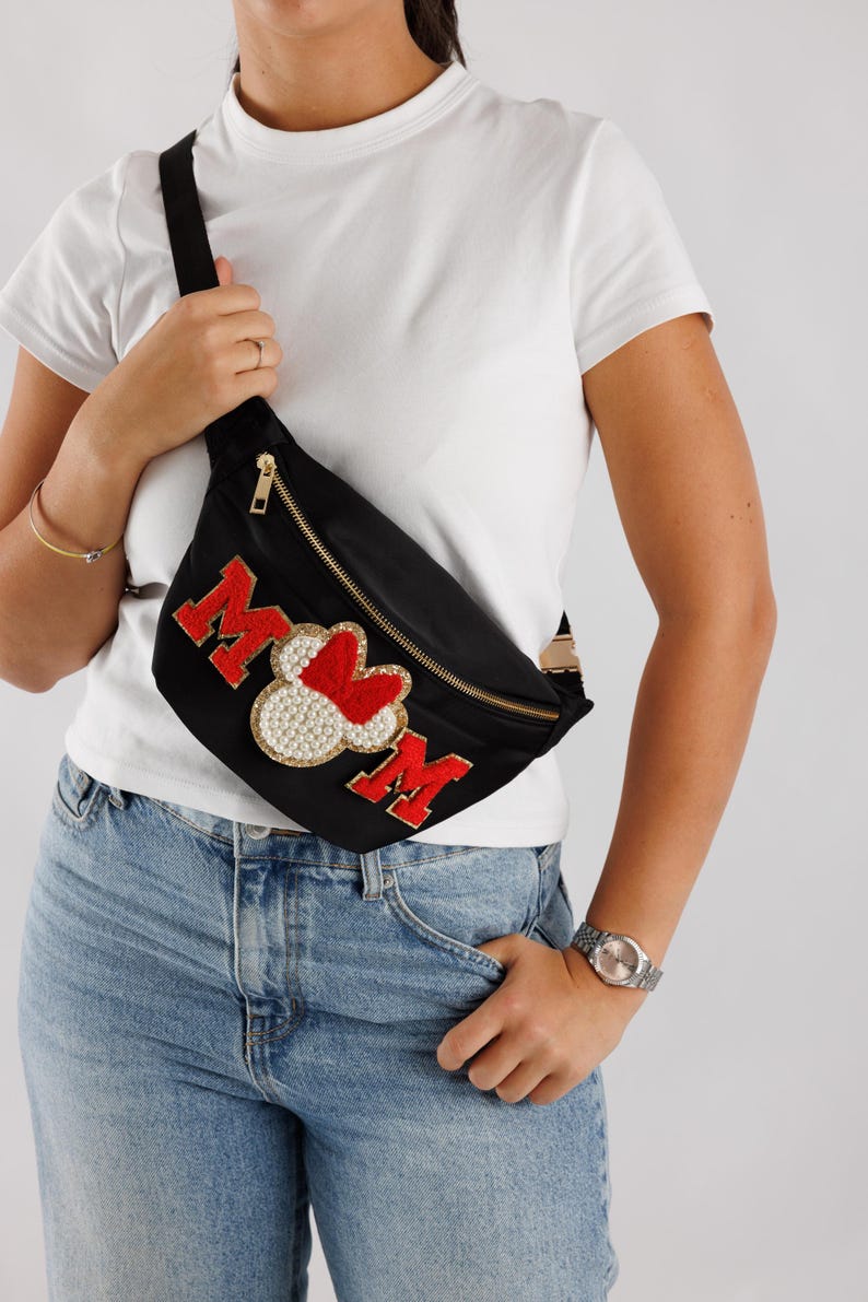 May include: Black fanny pack with gold zipper and adjustable strap. The pack features the word "MOM" in red, with a heart-shaped design and pearl accents. The bag is worn across the body.