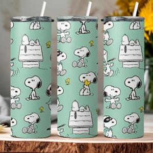 Puede incluir: Tres vasos de acero inoxidable con un fondo verde claro y un patrón repetido de Snoopy y su caseta. Los vasos tienen una pajita blanca y una tapa negra. El diseño incluye a Snoopy en varias poses y pájaros amarillos.