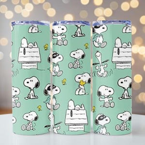 Puede incluir: Un juego de tres vasos cilíndricos con fondo verde claro que presenta un patrón repetitivo del personaje de dibujos animados Snoopy en varias poses, incluyendo sentado en una caseta de perro y con una capa de superhéroe, junto al personaje Woodstock.