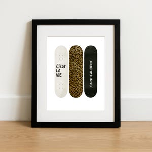 May include: Three skateboards in a black frame. One is white with "C'EST LA VIE" in black, the second has a leopard print, and the third is black with "SAINT LAURENT" in white. The artwork is displayed on a white background.