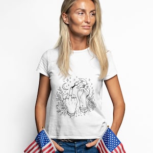 Könnte beinhalten: Weißes T-Shirt mit einer schwarz-weißen Grafik von Händen mit Schmuck, einem Halbmond und Sternen. Die Hände machen eine unhöfliche Geste. Die Person trägt eine blaue Jeans und hält zwei amerikanische Flaggen.