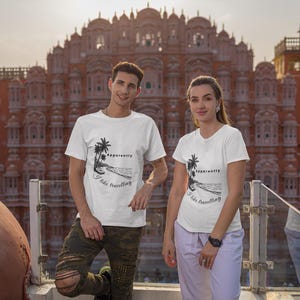 Könnte beinhalten: Zwei Personen tragen weiße T-Shirts mit einer schwarzen Grafik einer Palme, eines Strandes und dem Text "Apparently I like travelling". Der Hintergrund zeigt ein großes, verziertes, orangefarbenes Gebäude.