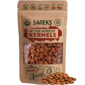 May include: A brown paper bag of bitter apricot kernels with a clear window, displaying the nuts inside. The bag has the text "SAREKS" and "BITTER APRICOT KERNELS". The bag also has USDA Organic and other health-related icons. A pile of kernels is in front of the bag.