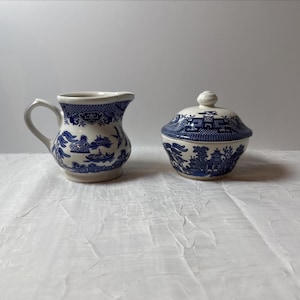 May include: A white and blue ceramic creamer and covered sugar bowl set. The blue design features a scenic landscape with buildings, trees, and water. The creamer has a handle and spout, while the sugar bowl has a lid with a small knob.